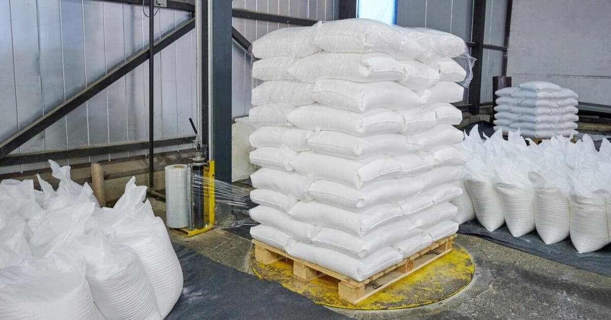 White sacks are stacked on a wood pallet beside a stretch wrap machine and plastic film rolls in a warehouse.