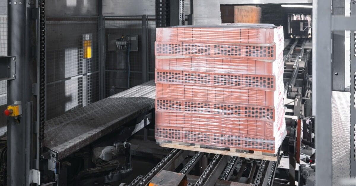 A pallet of orange bricks wrapped in clear plastic moves along a conveyor inside a gray manufacturing facility.