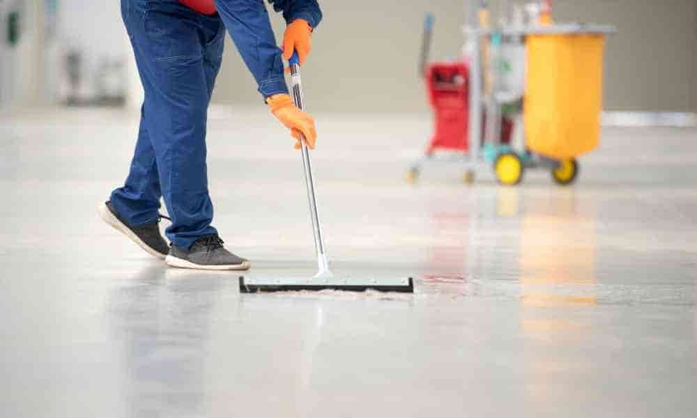  A worker wearing blue coveralls and orange gloves mops a floor. A commercial housekeeping cart stands in the background.