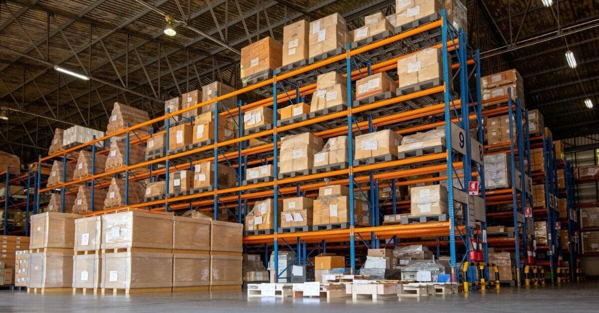A warehouse aisle with tall blue and orange racks holding cardboard boxes and wrapped pallets under high ceilings.