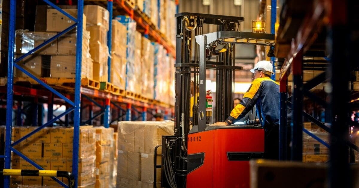 A worker in a white hard hat operating a red forklift between tall racks filled with cardboard boxes.