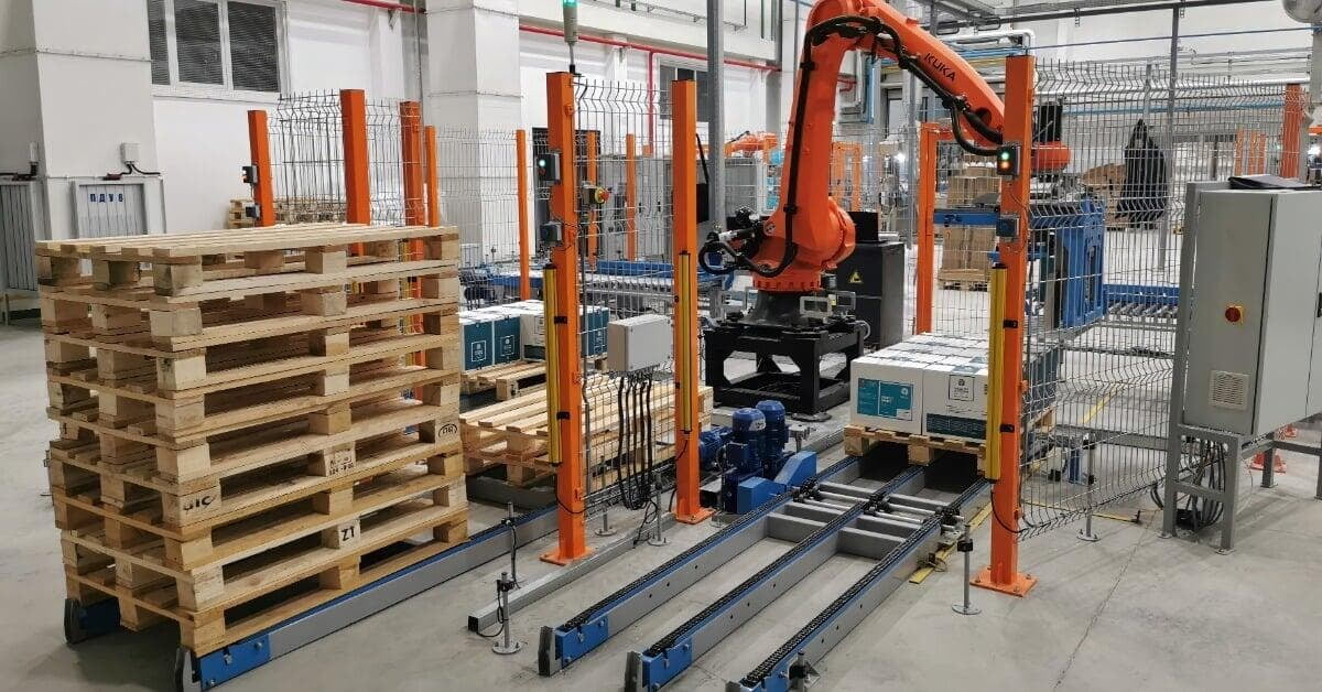 An orange robotic arm stacking boxes near wooden pallets, conveyors, and safety fencing inside a factory.