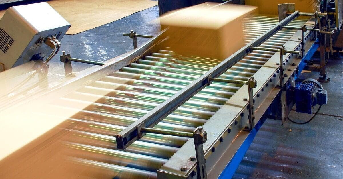 A cardboard box packing machine with a roller conveyor and metal housing inside a warehouse production area.