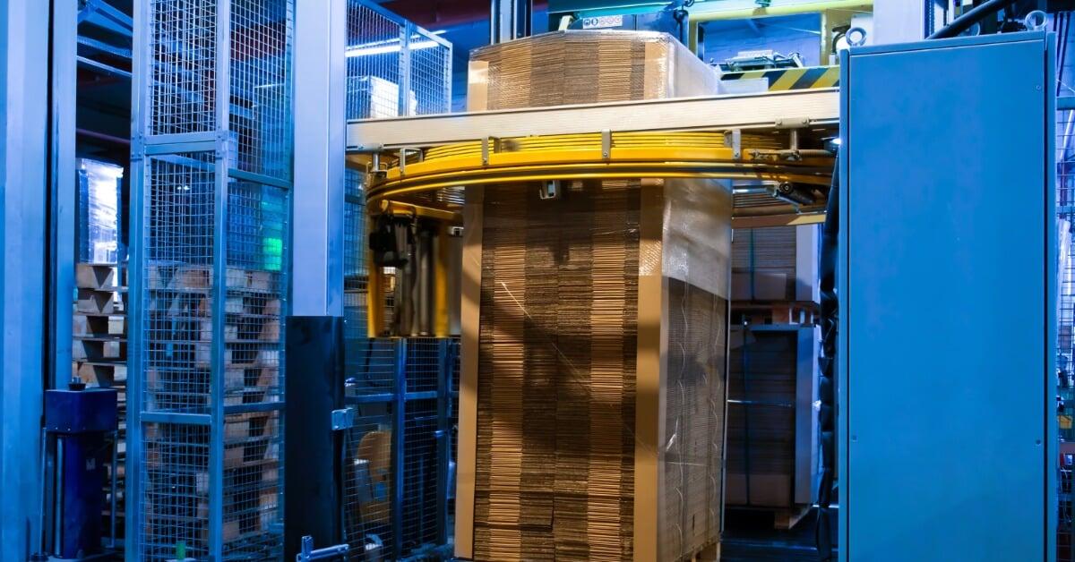 A pallet of stacked cardboard sits on a conveyor inside a stretch wrapping machine with yellow rails.