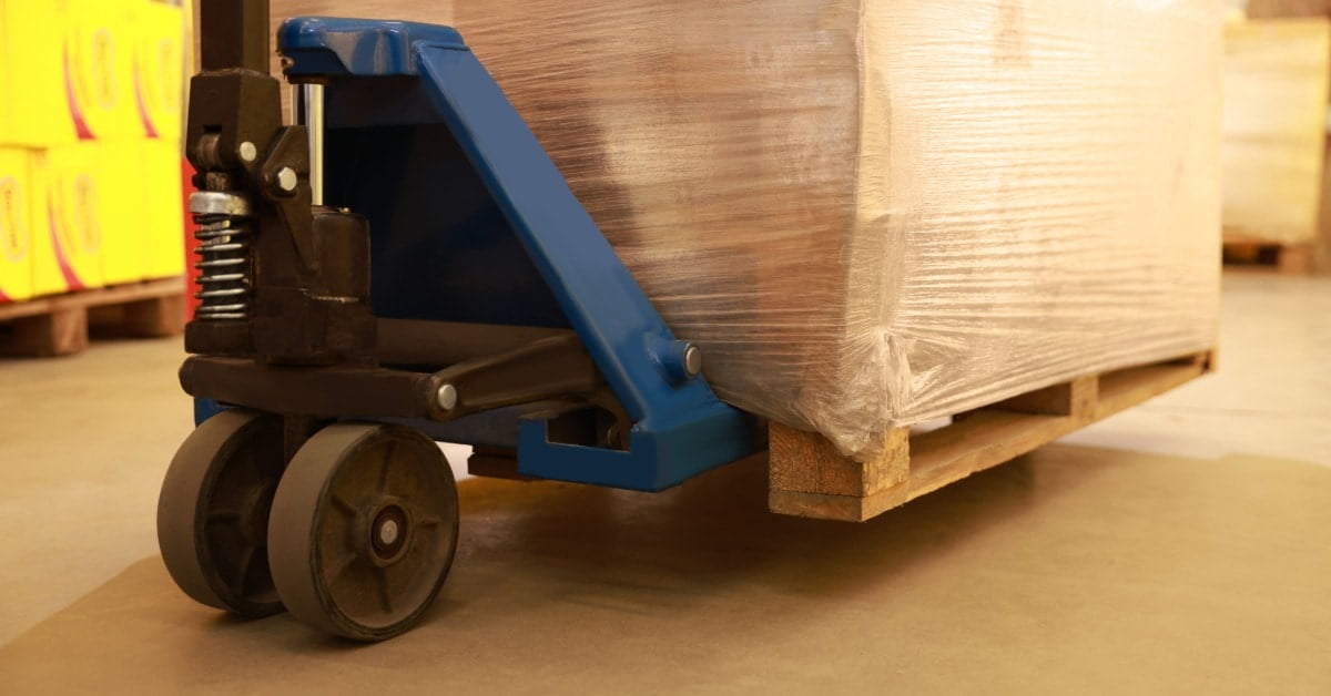 A blue pallet jack lifting a plastic-wrapped box on a wooden pallet inside a warehouse. Yellow boxes are in the background.
