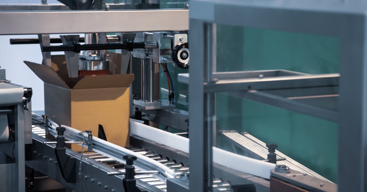 An open cardboard box sits under metal parts in a carton forming machine with rails and clear safety panels.
