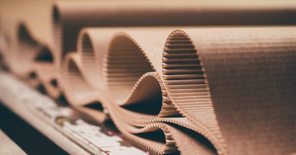 A close-up of curled corrugated cardboard sheets with brown ridges and folded edges. The sheets are on a processing line.