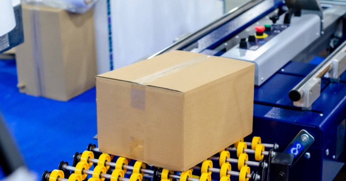 A sealed cardboard box moving on yellow rollers beside a blue carton sealing machine with buttons and metal rails.