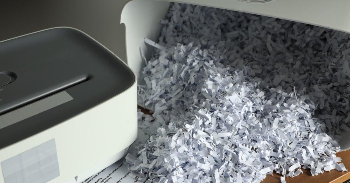 A white paper shredder with an open bin lying on its side is spilling shreds of paper across a wooden table.