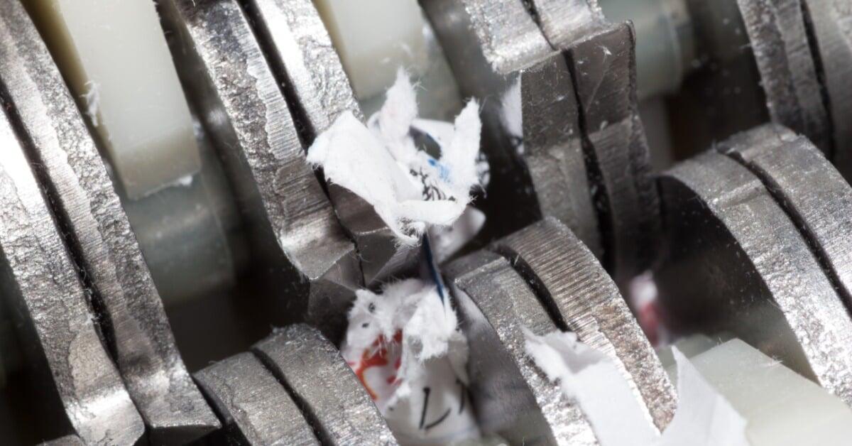 An extreme close-up of scraps of torn white paper caught between the teeth of metal paper shredder blades.