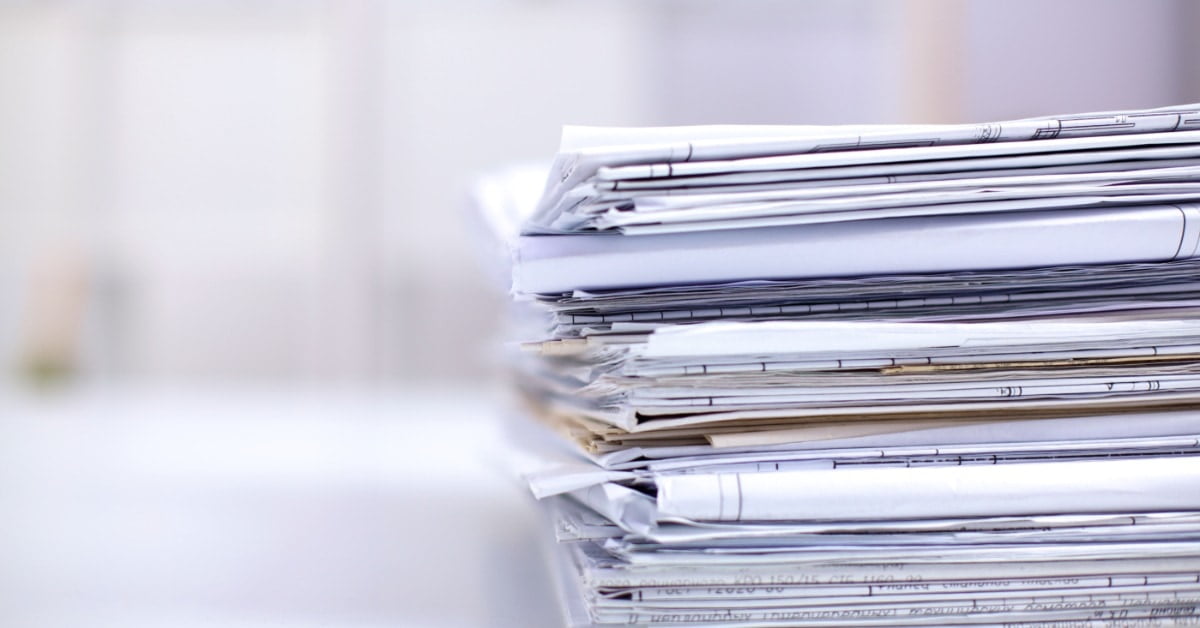 A tall stack of loose documents and folded papers rests on a desk. A light-colored room is blurred in the background.