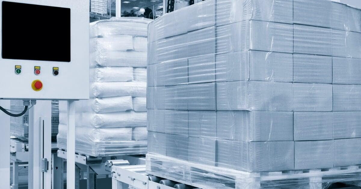 A plastic-wrapped pallet of stacked cardboard boxes sits on a roller conveyor beside a control panel in a warehouse.