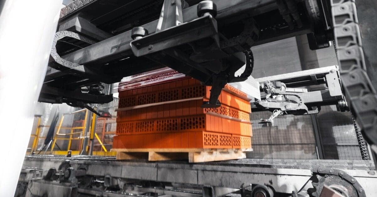 A pallet of orange bricks sits below overhead metal machinery on a conveyor. Wrapping film is on the left.