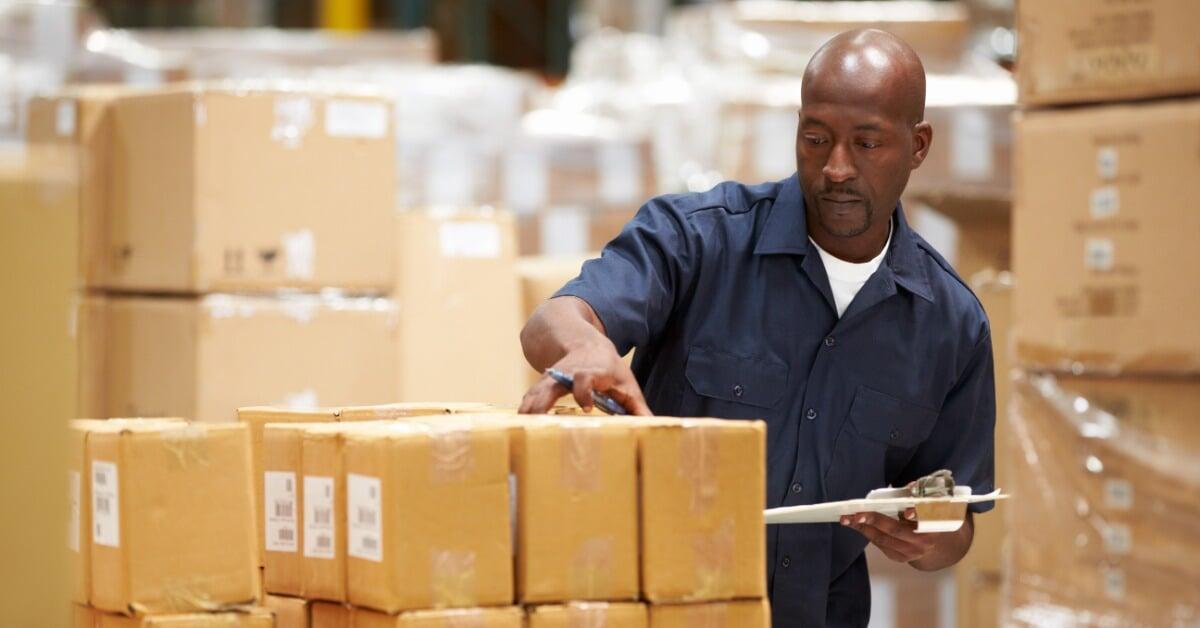 A man in a blue shirt holds a clipboard beside stacked cardboard boxes in a warehouse filled with packaged goods.
