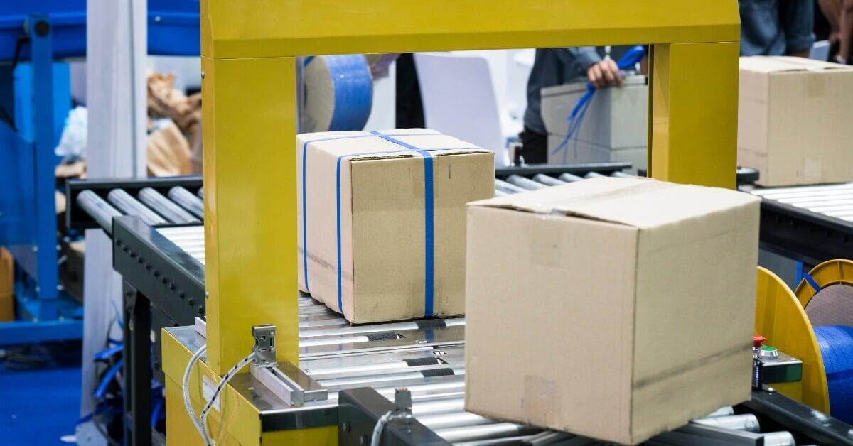 A cardboard box sits on conveyor rollers near a yellow strapping machine with a banded box behind it.