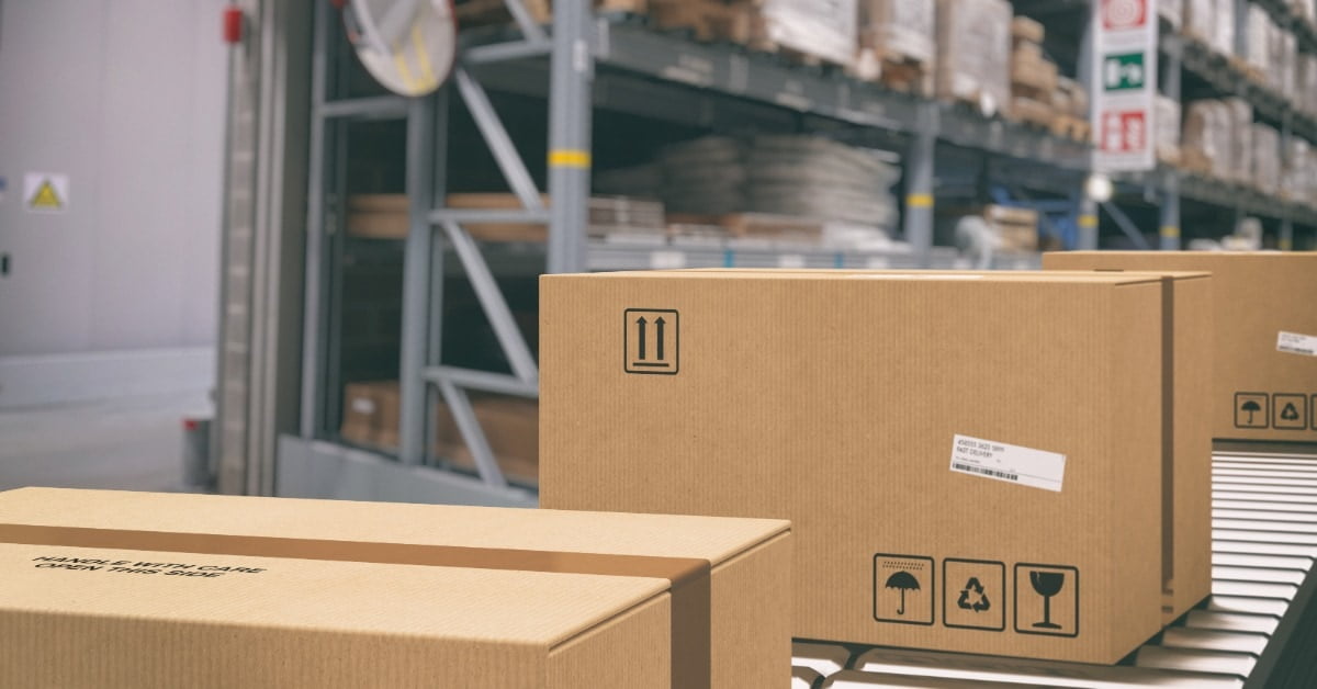 A group of cardboard boxes rests on conveyor rollers in a warehouse aisle with tall racks holding packaged goods.