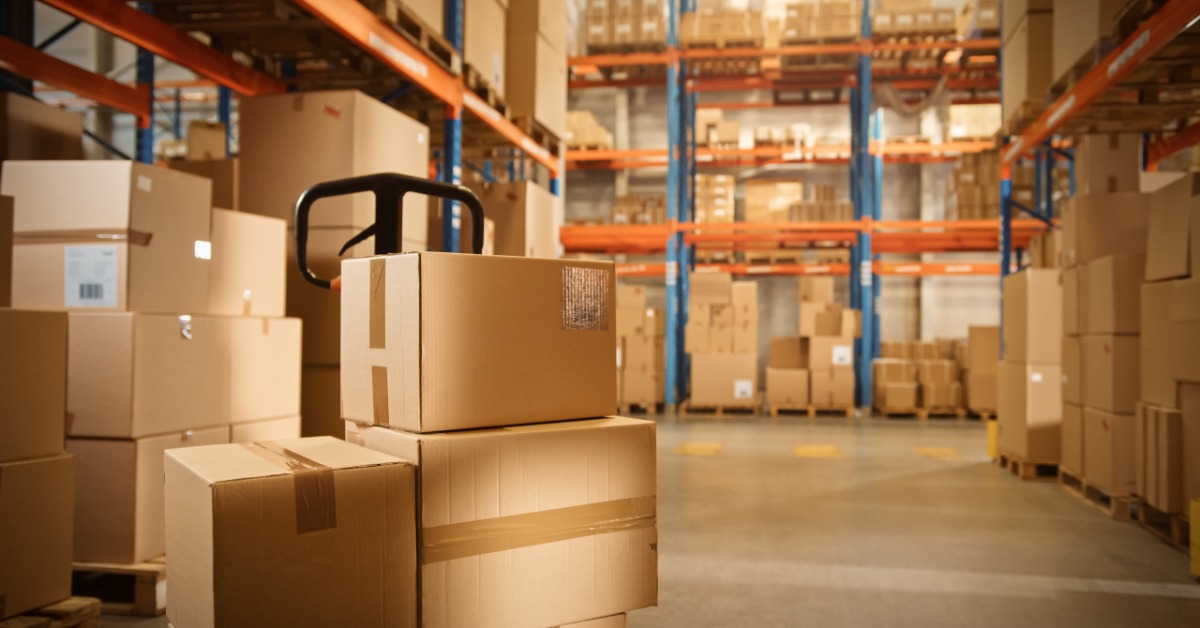 Cardboard boxes sit on a pallet truck in a warehouse aisle between tall shelves stacked with packages.