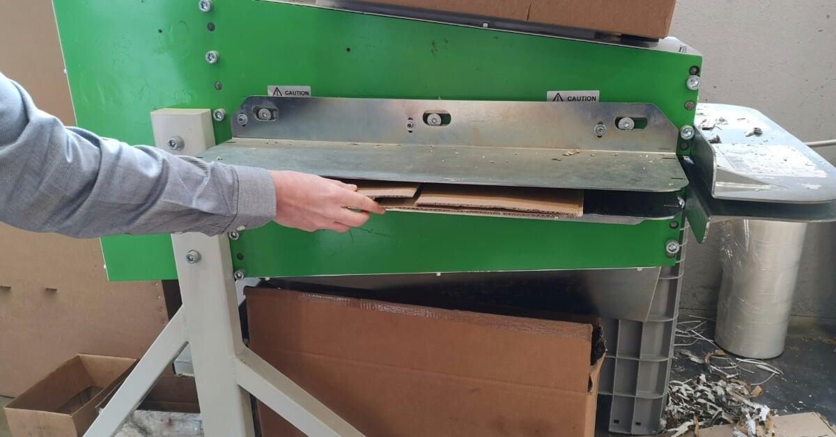 A hand feeding flat cardboard into a green shredder machine. Boxes and paper scraps lie on the floor.