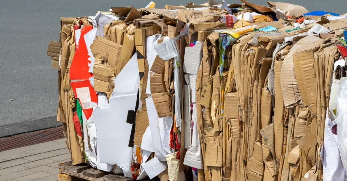 A large compressed bundle of flattened cardboard and paper resting on wooden pallets beside a paved surface.