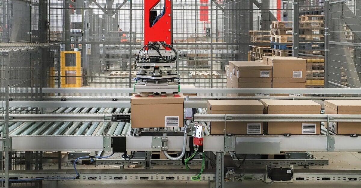 A red overhead palletizing machine lifting a cardboard box above metal roller conveyors inside a fenced warehouse line.
