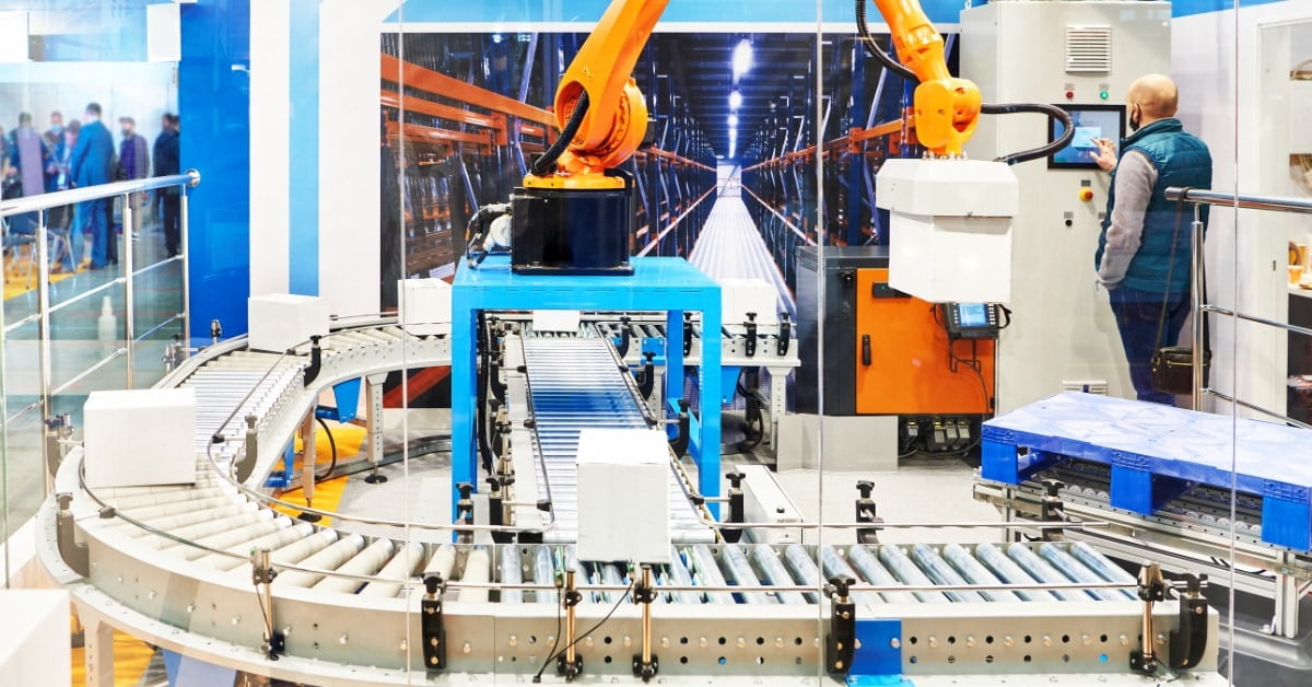 An orange industrial robotic arm above a curved roller conveyor with white boxes and a man at a control panel nearby.