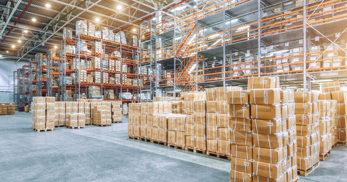 A large industrial warehouse with high racks filled with boxes, and cardboard boxes stacked on the floor in the foreground.