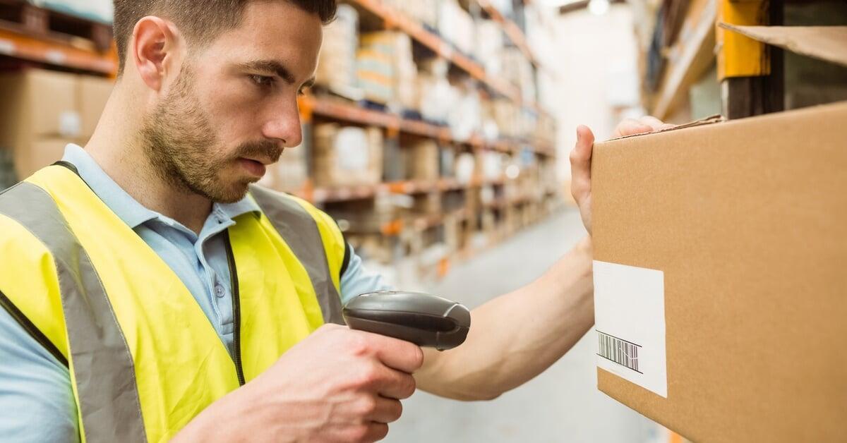 A male worker in a safety vest scans a barcode on a box in a warehouse aisle with shelves stacked with cardboard boxes.