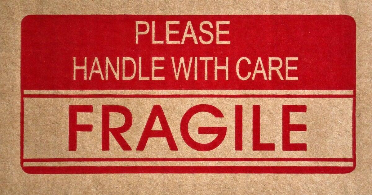 A brown cardboard box with a red labels that read 'FRAGILE' and 'PLEASE HANDLE WITH CARE' printed on the front of it.