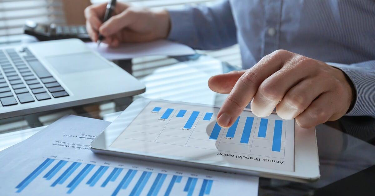 A businessman working at a desk with financial charts and a tablet displaying a bar graph, surrounded by documents.