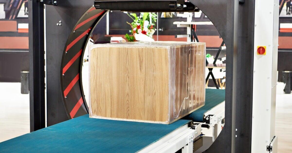 An orbital wrapper packing machine with a wooden box partially wrapped in clear plastic film on a blue conveyor belt.