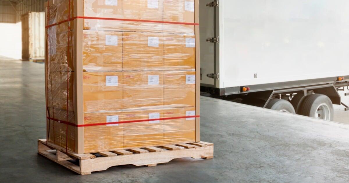 A stack of 12 boxes wrapped in plastic sits on a wooden pallet in an empty warehouse near a white semi-truck.