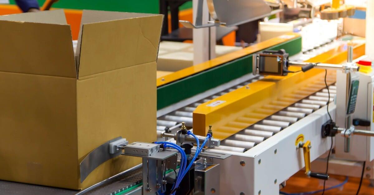 A cardboard box sits in front of the metal conveyor belt of a green and yellow automatic sealing machine.