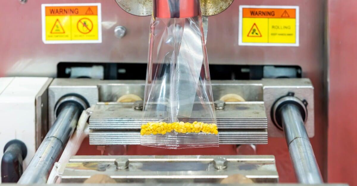 Small soybean granules fall into a plastic pouch held by a vertical automatic packing machine in a facility.