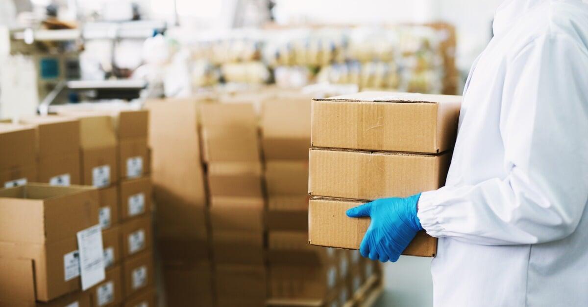 A person wearing blue robber gloves and a white jacket holds a stack of three cardboard boxes in front of a stack of boxes.