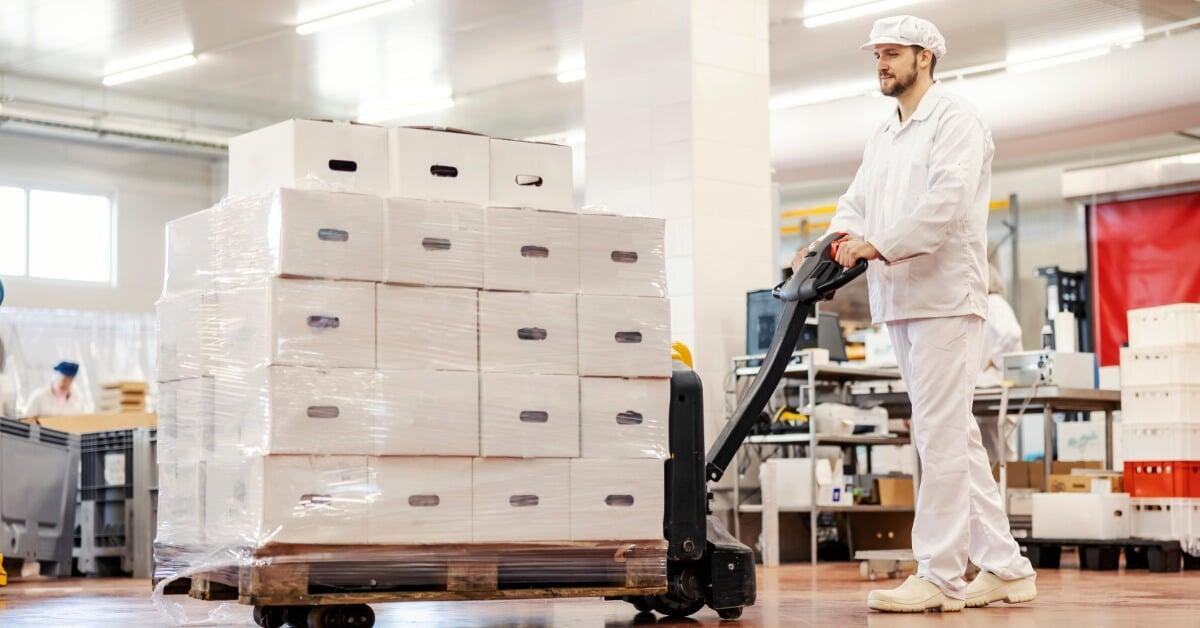 A factory worker dressed in white pushes a forklift that has a pallet with many white boxes stacked on it.