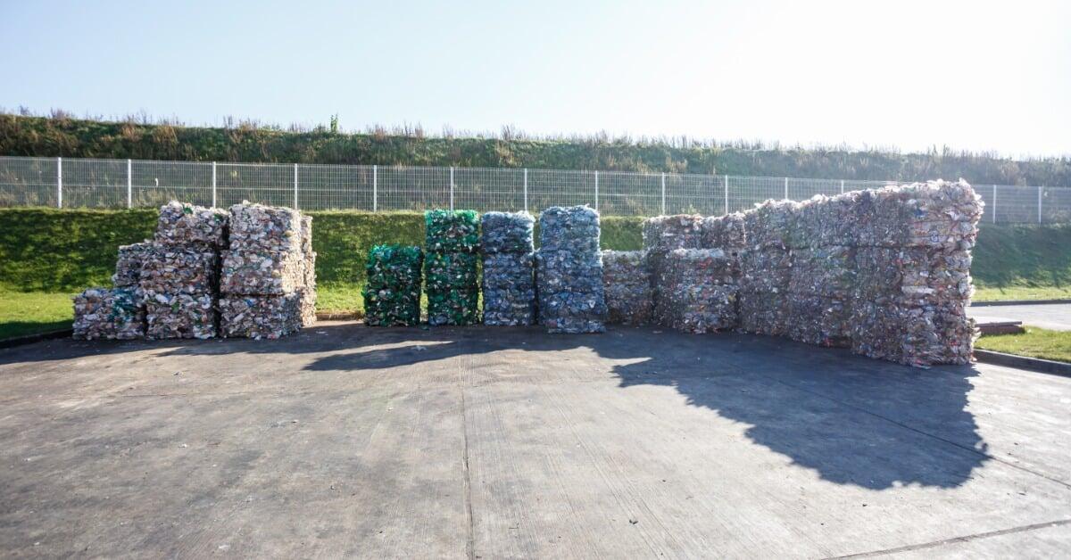 Multiple stacks of compressed plastic bales sit on a paved surface outdoors, arranged in rows with open space around them.