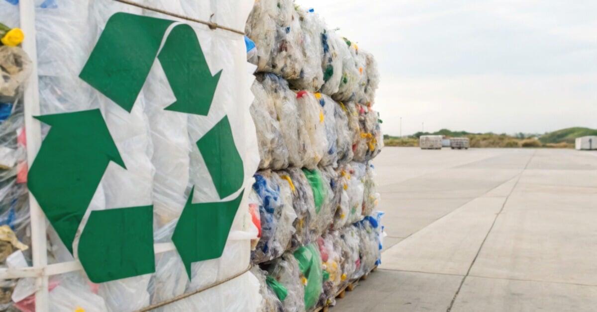 Large compressed plastic bales with a green recycling symbol are stacked outdoors on a concrete surface under an open sky.