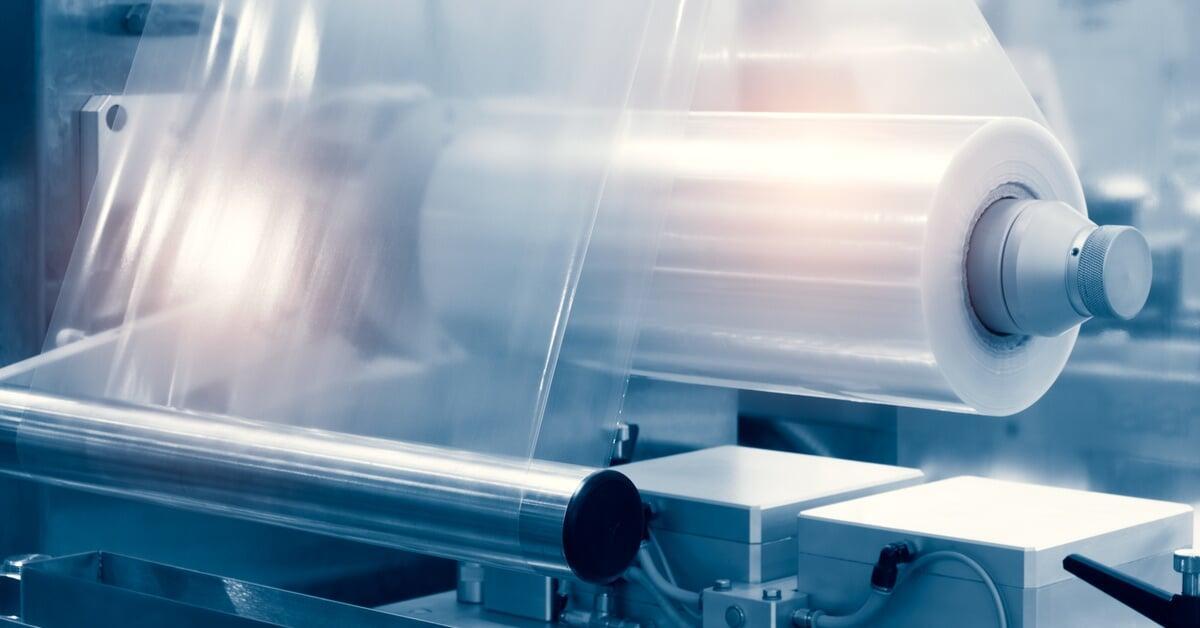 A roll of clear plastic film feeds through a production machine, with metal rollers and a blurred motion effect.