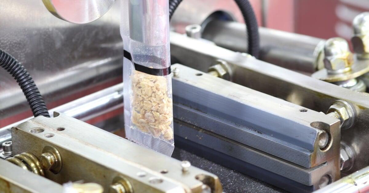 A vertical packaging machine fills a clear bag with dry beans, with metal parts and tubing visible around the process.