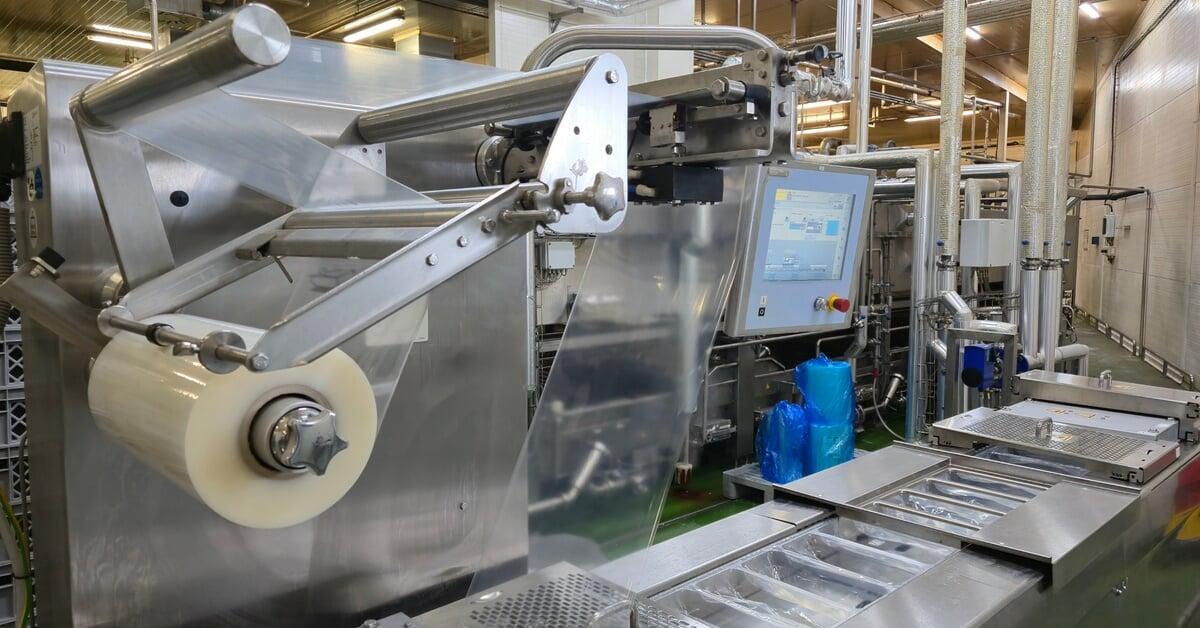 A large packaging machine with metal components, a control panel, and conveyor sections stands inside a factory setting.