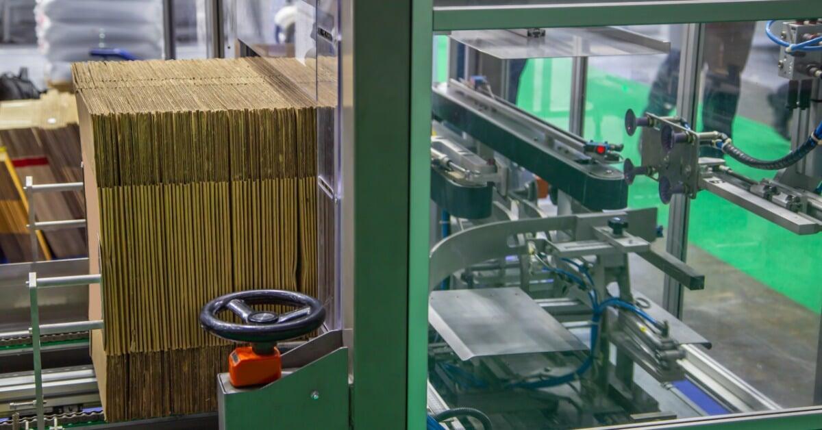 An automatic carton erector machine forms and opens flat cardboard boxes along a production line in a warehouse.