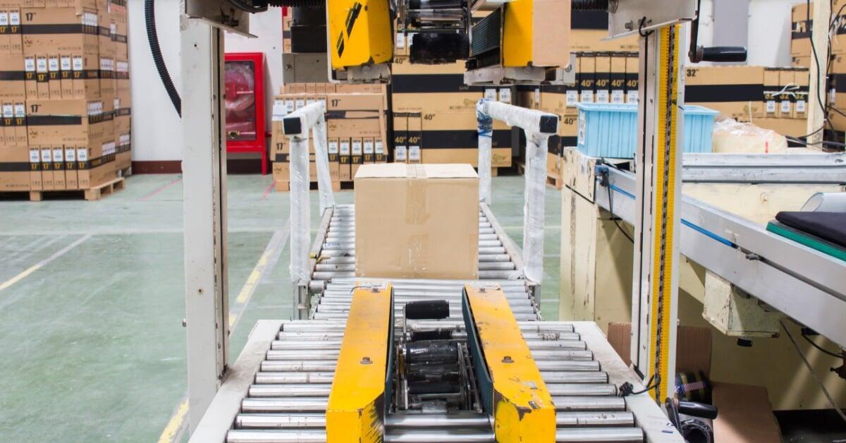 A box sits on a roller conveyor beneath a semi-automatic strapping machine, with stacked boxes stacked against a back wall.