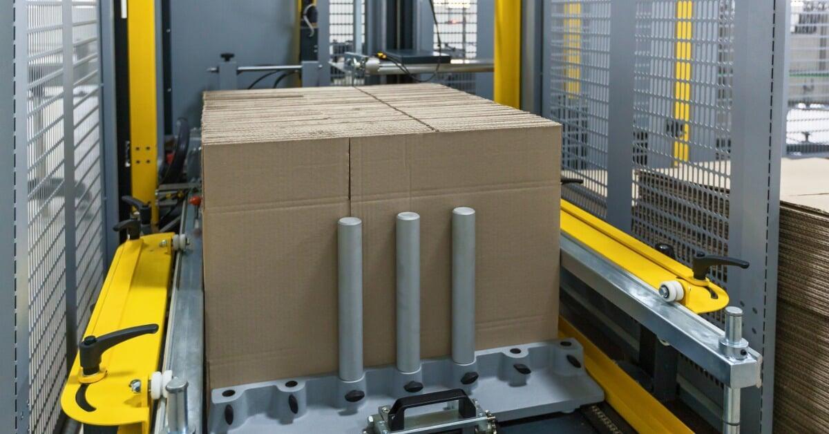 A cardboard box rests on a conveyor platform within a metal-framed strapping machine, surrounded by guard panels.