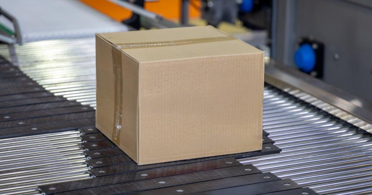 A sealed cardboard box moving along a conveyor belt with rollers and packaging machinery in the background.