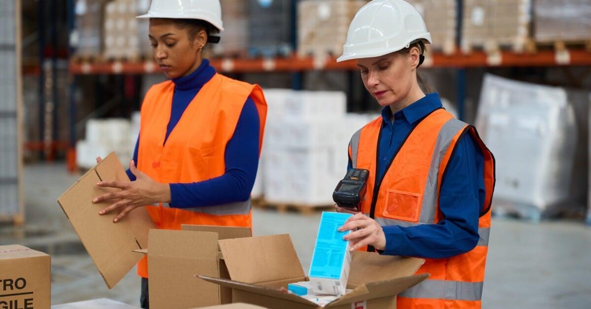 A pair of women packing cardboard boxes at a warehouse table with shelves, carts, and parcels nearby.