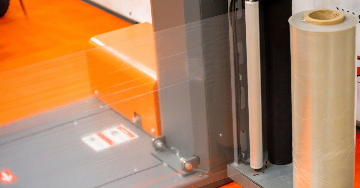 A close-up of a large roll of clear plastic film mounted on a piece of orange industrial production equipment.