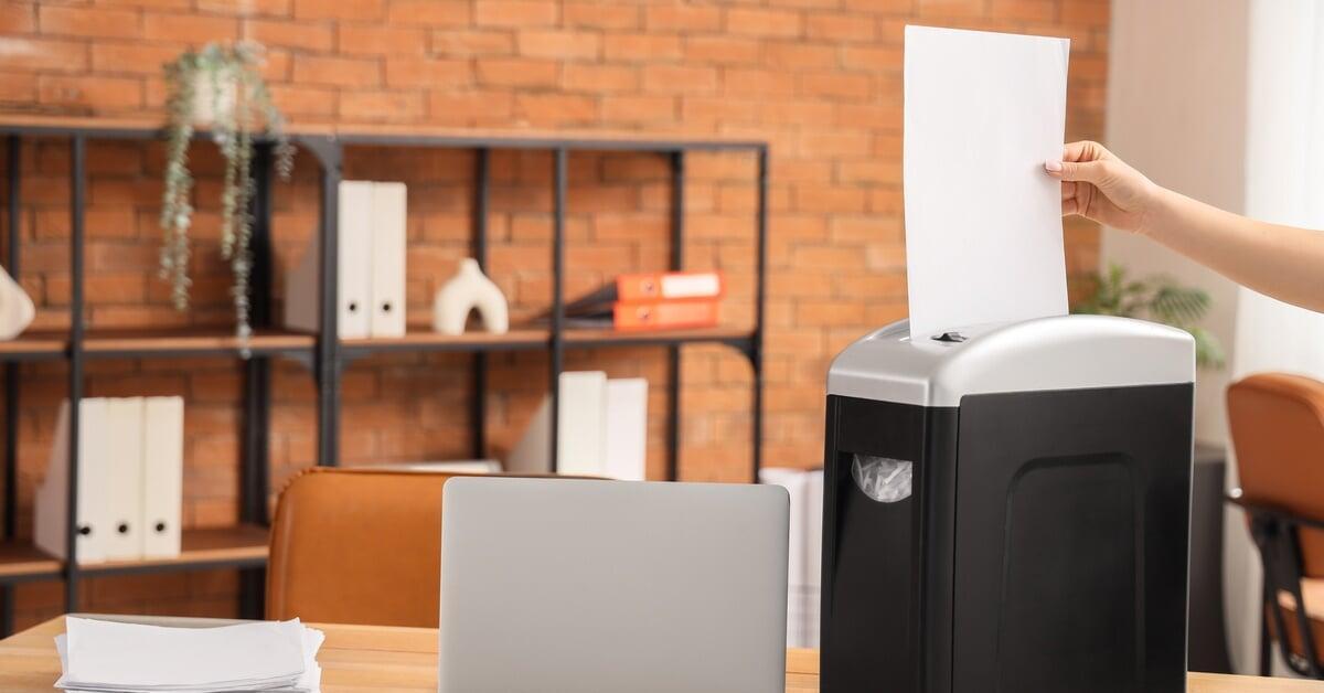 A hand feeding a white paper sheet into a black shredder on a desk near office items on a light desktop surface.