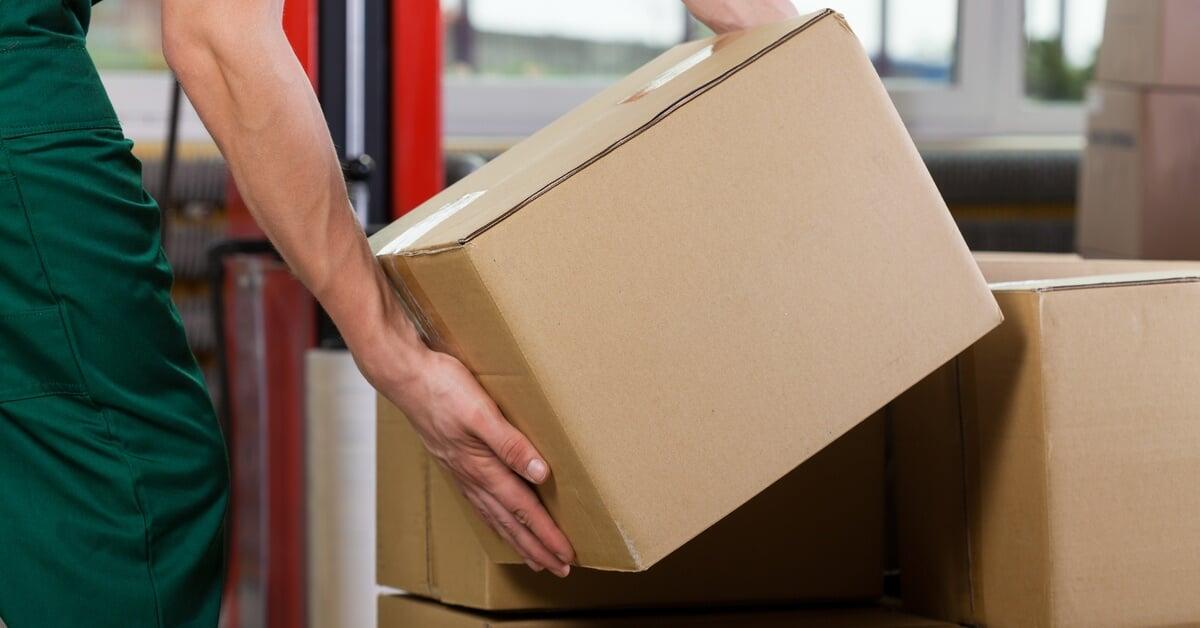 A warehouse worker wearing gloves lifts a cardboard box, with storage shelves and packaged items visible in the background.