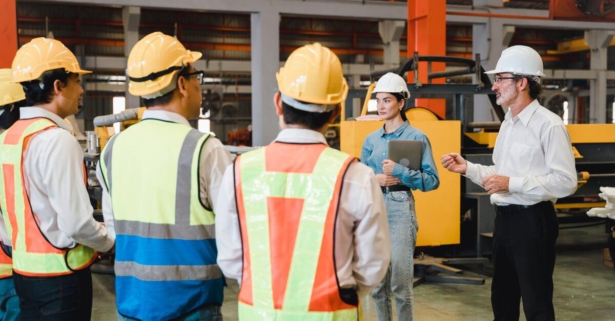 A man and woman in hard hats stand on a factory floor in front of four people in hard hats and reflective vests.