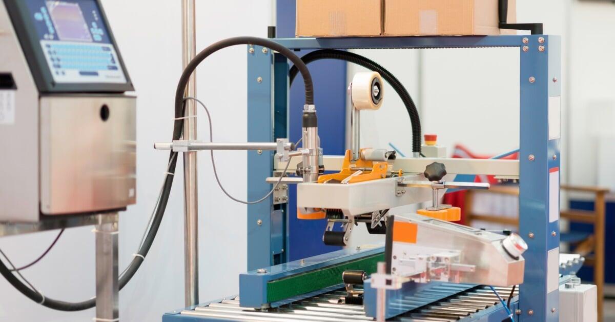 A carton sealing machine and conveyor belt moving a cardboard box through an industrial packing line.